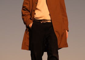 Young man in a brown coat poses in a grassy field at sunset, exuding elegance and style.